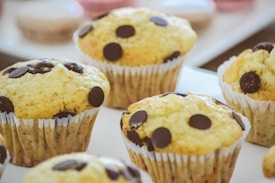 A close-up view of several freshly baked muffins with chocolate chips on top. The muffins are placed in white paper liners and are arranged on a light-colored surface. In the blurred background, there are additional baked goods, possibly macarons.