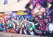 A vibrant and colorful graffiti mural on a wall. The artwork involves abstract shapes and bold text with phrases like 'GLOBAL DOMINATION'. The right side of the mural features a larger-than-life depiction of a robotic or armored figure in purples and pinks. The background includes swirling patterns resembling galaxies and cosmic scenes, with vivid blues, oranges, and purples creating a dynamic visual impact.
