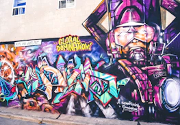 A vibrant and colorful graffiti mural on a wall. The artwork involves abstract shapes and bold text with phrases like 'GLOBAL DOMINATION'. The right side of the mural features a larger-than-life depiction of a robotic or armored figure in purples and pinks. The background includes swirling patterns resembling galaxies and cosmic scenes, with vivid blues, oranges, and purples creating a dynamic visual impact.