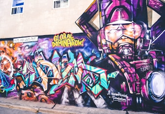 A vibrant and colorful graffiti mural on a wall. The artwork involves abstract shapes and bold text with phrases like 'GLOBAL DOMINATION'. The right side of the mural features a larger-than-life depiction of a robotic or armored figure in purples and pinks. The background includes swirling patterns resembling galaxies and cosmic scenes, with vivid blues, oranges, and purples creating a dynamic visual impact.