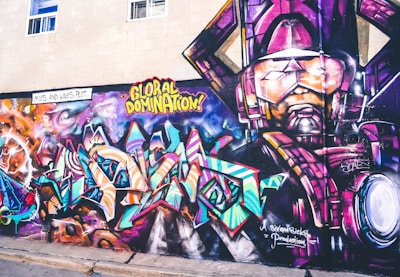A vibrant and colorful graffiti mural on a wall. The artwork involves abstract shapes and bold text with phrases like 'GLOBAL DOMINATION'. The right side of the mural features a larger-than-life depiction of a robotic or armored figure in purples and pinks. The background includes swirling patterns resembling galaxies and cosmic scenes, with vivid blues, oranges, and purples creating a dynamic visual impact.