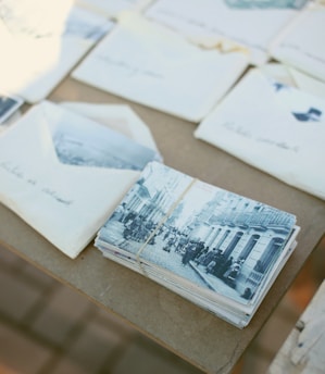 A stack of vintage photographs and letters bound with a rubber band rests on a table. The photographs depict a busy street scene with people walking along a cobblestone road lined with old buildings. Several envelopes, some partially open, are scattered around the stack, suggesting a collection of personal correspondence.