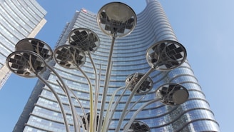 A futuristic sculpture stands in front of a tall, modern glass skyscraper with a curved facade. The sculpture resembles a cluster of tall, slender stems with round, solar-panel-like discs at the top. The sky is clear and blue, enhancing the urban and contemporary atmosphere.