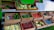 An open market stall displays a variety of fresh produce in wooden crates. Vegetables such as cabbage, carrots, beets, cucumbers, tomatoes, and green beans are arranged neatly. Small trays hold potatoes, berries, and other colorful fruits. A menu board listing meat and eggs is visible in the background.
