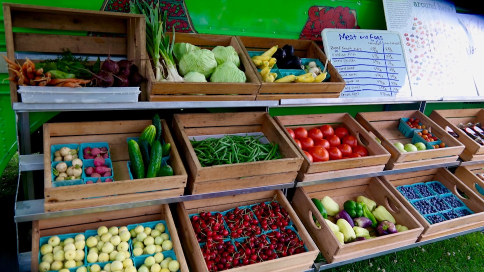 An open market stall displays a variety of fresh produce in wooden crates. Vegetables such as cabbage, carrots, beets, cucumbers, tomatoes, and green beans are arranged neatly. Small trays hold potatoes, berries, and other colorful fruits. A menu board listing meat and eggs is visible in the background.