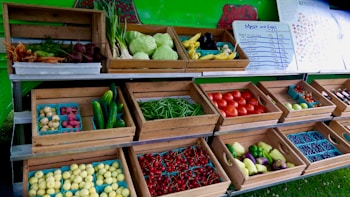 An open market stall displays a variety of fresh produce in wooden crates. Vegetables such as cabbage, carrots, beets, cucumbers, tomatoes, and green beans are arranged neatly. Small trays hold potatoes, berries, and other colorful fruits. A menu board listing meat and eggs is visible in the background.