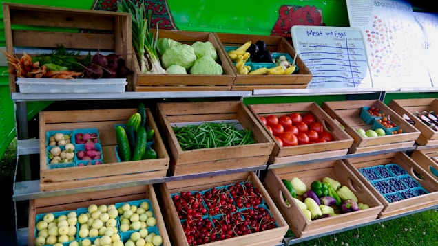 An open market stall displays a variety of fresh produce in wooden crates. Vegetables such as cabbage, carrots, beets, cucumbers, tomatoes, and green beans are arranged neatly. Small trays hold potatoes, berries, and other colorful fruits. A menu board listing meat and eggs is visible in the background.