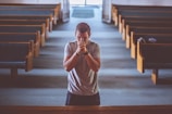 A man in a gray shirt stands in the central aisle of an empty church, with wooden pews on either side. He appears to be praying, with his hands clasped in front of his face and eyes closed. The lighting is soft, coming from large windows at the far end, creating a peaceful and introspective atmosphere.