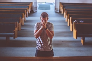 A man in a gray shirt stands in the central aisle of an empty church, with wooden pews on either side. He appears to be praying, with his hands clasped in front of his face and eyes closed. The lighting is soft, coming from large windows at the far end, creating a peaceful and introspective atmosphere.