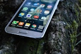A smartphone with a variety of apps displaying on its screen is resting on a textured outdoor surface, possibly a mossy log. The apps visible include WhatsApp, YouTube, and Instagram, among others.
