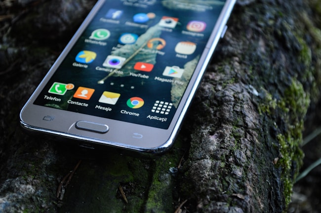 A smartphone with a variety of apps displaying on its screen is resting on a textured outdoor surface, possibly a mossy log. The apps visible include WhatsApp, YouTube, and Instagram, among others.
