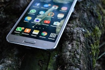 A smartphone with a variety of apps displaying on its screen is resting on a textured outdoor surface, possibly a mossy log. The apps visible include WhatsApp, YouTube, and Instagram, among others.