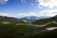 A scenic view of a winding road cutting through lush green hills with mountains in the background under a partly cloudy blue sky. The landscape is expansive and vibrant, offering a sense of tranquility. Several vehicles are seen traveling along the road.