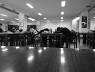 People are seated at wooden tables in a room that appears to be a library or study area. Most of them have their heads down on the tables, possibly resting or sleeping. The room is lit with overhead fluorescent lights and has bookshelves and framed art on the walls.