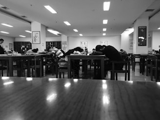 People are seated at wooden tables in a room that appears to be a library or study area. Most of them have their heads down on the tables, possibly resting or sleeping. The room is lit with overhead fluorescent lights and has bookshelves and framed art on the walls.