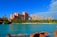 A large, pink-toned resort complex stands prominently against a clear blue sky. The architecture features multiple towers connected by a central bridge structure. Lush greenery and palm trees surround the buildings, and a tranquil blue lagoon is in the foreground, bordered by rocky, reddish terrain.