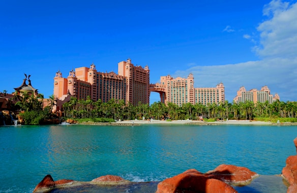 A large, pink-toned resort complex stands prominently against a clear blue sky. The architecture features multiple towers connected by a central bridge structure. Lush greenery and palm trees surround the buildings, and a tranquil blue lagoon is in the foreground, bordered by rocky, reddish terrain.