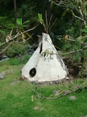A traditional teepee made of canvas is set up outdoors surrounded by lush greenery. The structure has a circular opening as its entrance, and wooden poles extend upwards from its apex. The ground is covered with grass and there are scattered rocks around the teepee.
