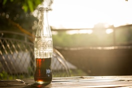 A glass soda bottle containing a dark liquid is placed on a wooden surface with blurred greenery in the background. Sunlight creates a warm glow around the bottle, casting shadows on the surface.