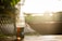 A glass soda bottle containing a dark liquid is placed on a wooden surface with blurred greenery in the background. Sunlight creates a warm glow around the bottle, casting shadows on the surface.