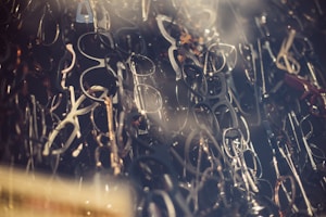 A dense collection of various eyeglass frames overlapping with each other, creating an intricate and somewhat chaotic visual pattern. The frames, in different shapes and sizes, are primarily black, brown, and metallic, and are stacked closely together.