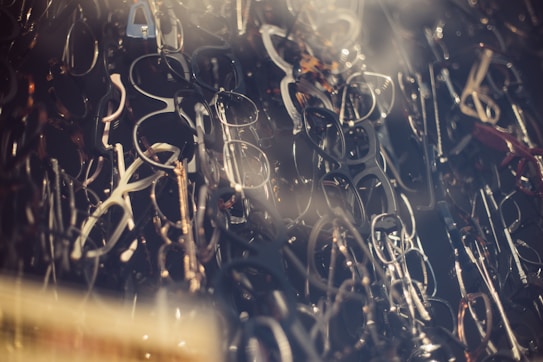 A dense collection of various eyeglass frames overlapping with each other, creating an intricate and somewhat chaotic visual pattern. The frames, in different shapes and sizes, are primarily black, brown, and metallic, and are stacked closely together.