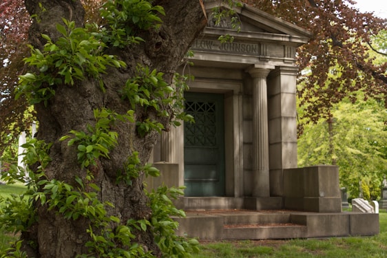 A large tree with lush green leaves partially obscures a stone mausoleum with classic columns and an engraved name at the top. The structure is set in a verdant area with additional trees and gravestones visible in the background.