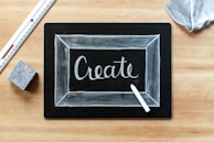 A blackboard with the word 'Create' written in white chalk, surrounded by a thin chalk-drawn border. A piece of white chalk and a ruler are placed on a wooden surface, alongside an eraser and a piece of fabric.