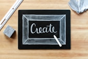 A blackboard with the word 'Create' written in white chalk, surrounded by a thin chalk-drawn border. A piece of white chalk and a ruler are placed on a wooden surface, alongside an eraser and a piece of fabric.