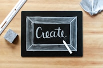 A blackboard with the word 'Create' written in white chalk, surrounded by a thin chalk-drawn border. A piece of white chalk and a ruler are placed on a wooden surface, alongside an eraser and a piece of fabric.