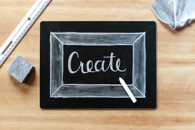A blackboard with the word 'Create' written in white chalk, surrounded by a thin chalk-drawn border. A piece of white chalk and a ruler are placed on a wooden surface, alongside an eraser and a piece of fabric.