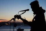 A silhouette of a musician playing the guitar and singing into a microphone set against a twilight sky. The background features a serene body of water and distant city lights, creating a peaceful ambiance.