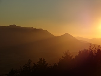 A serene mountain landscape during a golden sunset. Silhouettes of mountains create a dramatic horizon against a glowing sky, with the sun casting warm, golden light over the scene. Foreground trees are visible in darkness, adding depth to the image.