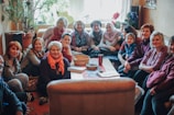 A group of people, mostly women, sit closely together in a cozy room, smiling warmly. The room is filled with natural light and decorated with plants and flowers. On the table in front of them is a basket, a cup, and some notebooks.