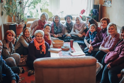 A group of people, mostly women, sit closely together in a cozy room, smiling warmly. The room is filled with natural light and decorated with plants and flowers. On the table in front of them is a basket, a cup, and some notebooks.