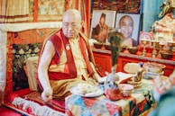 A Buddhist monk wearing traditional robes sits in an ornately decorated room. The setting includes vibrant textiles and intricate patterns, with numerous religious artifacts and photographs arranged thoughtfully. The atmosphere is serene and reflective.