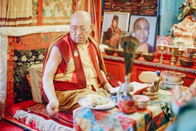 A Buddhist monk wearing traditional robes sits in an ornately decorated room. The setting includes vibrant textiles and intricate patterns, with numerous religious artifacts and photographs arranged thoughtfully. The atmosphere is serene and reflective.