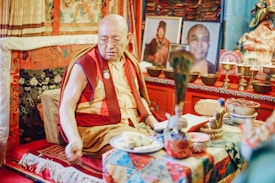 A Buddhist monk wearing traditional robes sits in an ornately decorated room. The setting includes vibrant textiles and intricate patterns, with numerous religious artifacts and photographs arranged thoughtfully. The atmosphere is serene and reflective.