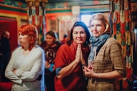 A group of women in a colorful indoor setting with ornate and patterned decorations. The focus is on three women, with one making a gesture with her hands in front, while the others stand beside and behind her. The background is out of focus, adding to the intimate and warm ambiance.