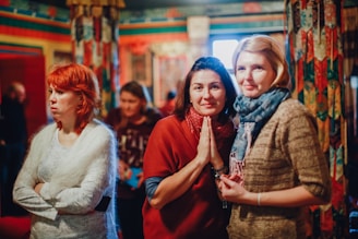 A group of women in a colorful indoor setting with ornate and patterned decorations. The focus is on three women, with one making a gesture with her hands in front, while the others stand beside and behind her. The background is out of focus, adding to the intimate and warm ambiance.