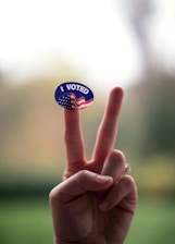 A hand gestures with two fingers raised, resembling a 'V' sign. A sticker with the text 'I VOTED' and an American flag design is placed on one of the fingers. The background is softly blurred.