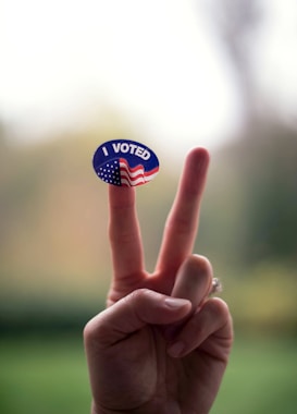 A hand gestures with two fingers raised, resembling a 'V' sign. A sticker with the text 'I VOTED' and an American flag design is placed on one of the fingers. The background is softly blurred.