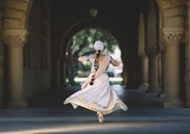 A person dressed in traditional Indian attire is dancing in a shaded stone archway. The bright garment features elaborate patterns and a mix of gold, white, and green colors. The dancer's hair is adorned with flowers, and the setting suggests an outdoor structure, possibly a historical or cultural site with architectural columns on each side.