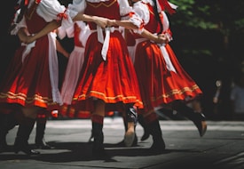 A group of people in traditional, vibrant red and white costumes are dancing on stage. The dancers are arranged in a circle, holding hands, and mid-movement, creating a dynamic sense of motion. The costumes have intricate designs and patterns, featuring white shirts with red vests and skirts decorated with golden embroidery. They wear black boots, and the lighting creates a lively yet slightly shadowed atmosphere.