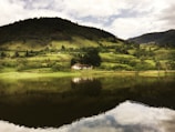 A tranquil rural landscape featuring a small, isolated house nestled at the base of a lush, green hill. The scene reflects in a still body of water in the foreground, creating a mirror-like effect. The sky is partially cloudy, casting soft light across the rolling hills and dense vegetation.