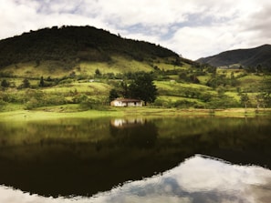 A tranquil rural landscape featuring a small, isolated house nestled at the base of a lush, green hill. The scene reflects in a still body of water in the foreground, creating a mirror-like effect. The sky is partially cloudy, casting soft light across the rolling hills and dense vegetation.