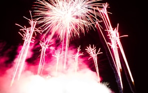 Brilliant bursts of fireworks light up a dark sky, with vibrant streaks of red and white emanating from the explosion. Smoke from the pyrotechnics creates a cloud-like effect, adding depth to the scene.