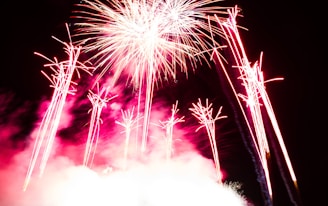 Brilliant bursts of fireworks light up a dark sky, with vibrant streaks of red and white emanating from the explosion. Smoke from the pyrotechnics creates a cloud-like effect, adding depth to the scene.