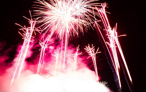 Brilliant bursts of fireworks light up a dark sky, with vibrant streaks of red and white emanating from the explosion. Smoke from the pyrotechnics creates a cloud-like effect, adding depth to the scene.