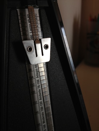 A traditional mechanical metronome with a metal pendulum and a scale marked with tempo ranges. The device is black with silver metallic components.